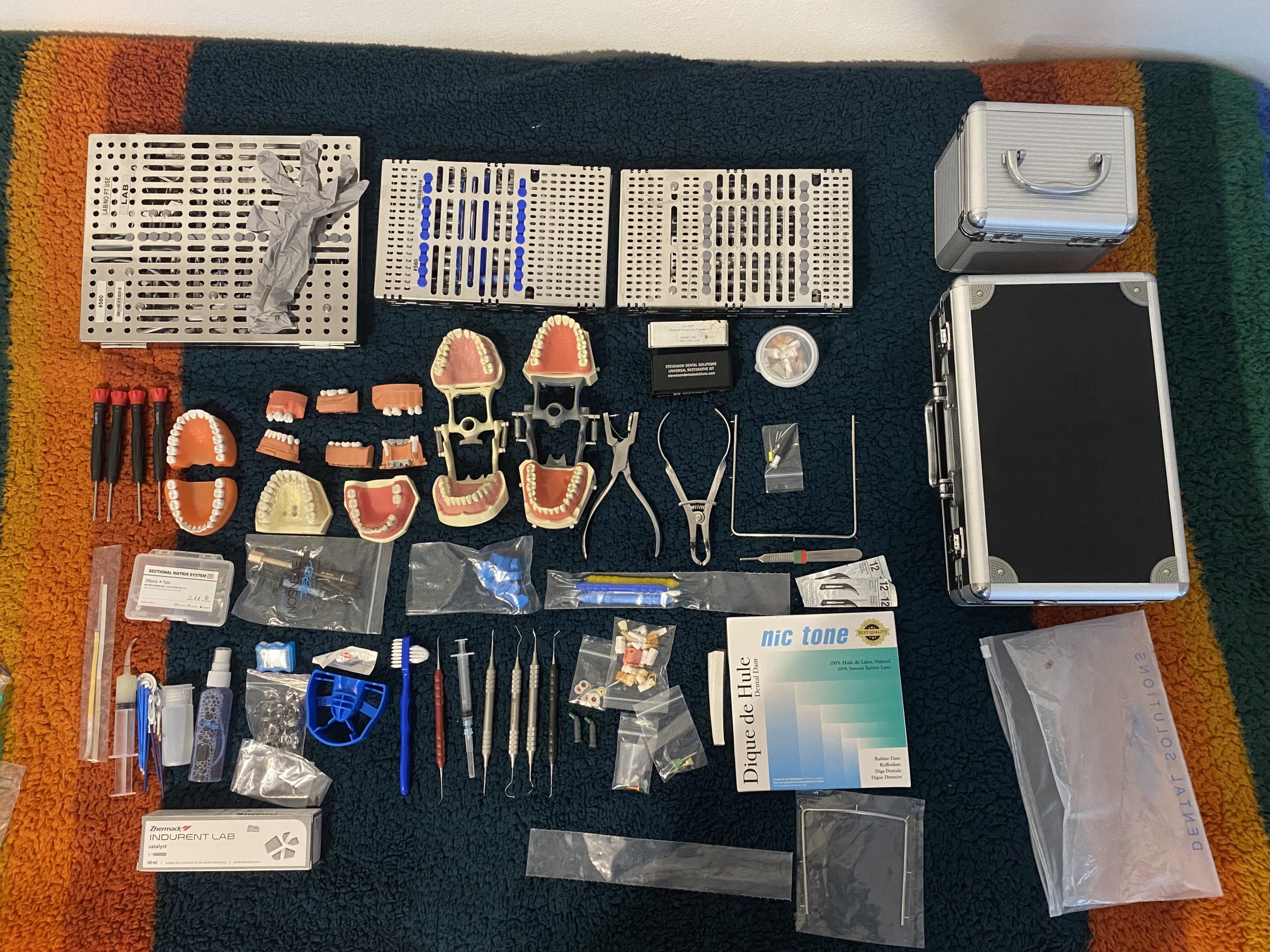 毛布の上に並べられた歯科用器具一式。滅菌カセット3枚、歯列模型、鉗子、ラバーダムシート、印象材、アルミケース2個などが写っている