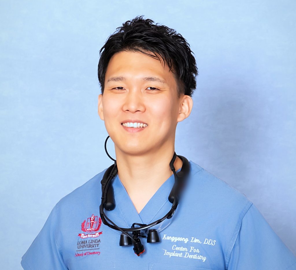 青い術衣に拡大鏡をかけ、正面を向いて微笑む歯科医師 林剛永(Kangyeong Lim, DDS)