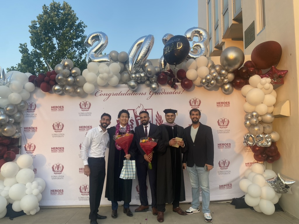 Loma Linda University の卒業式当日、記念撮影用のバックドロップ前で、ガウンとレイを身につけた著者が同期とともに花束を手に立っている