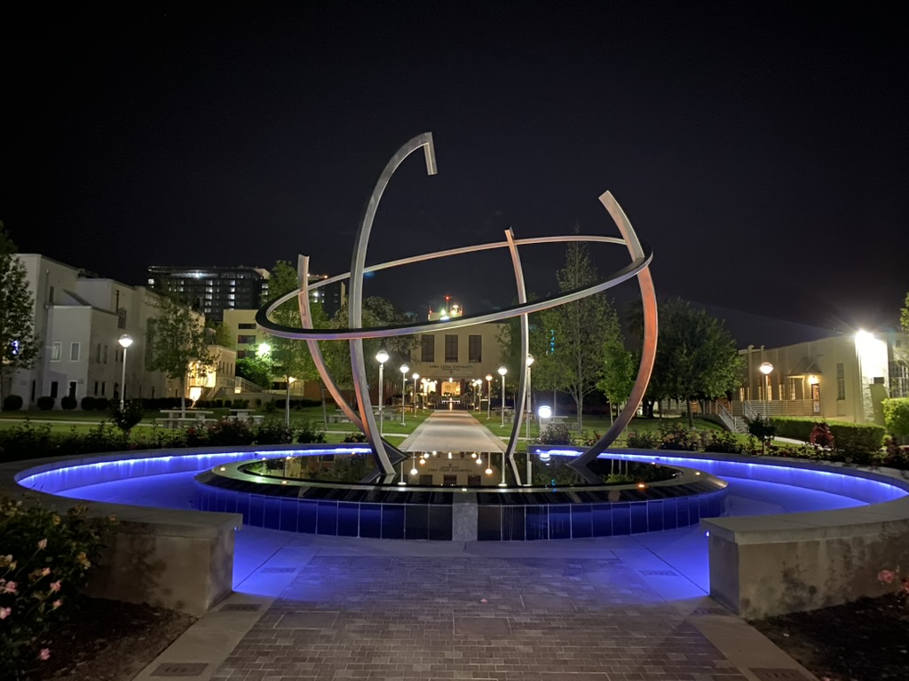 夜の Loma Linda University キャンパス。円形の噴水と金属製の球体モニュメントの奥に、Loma Linda University と掲げられた建物が見える