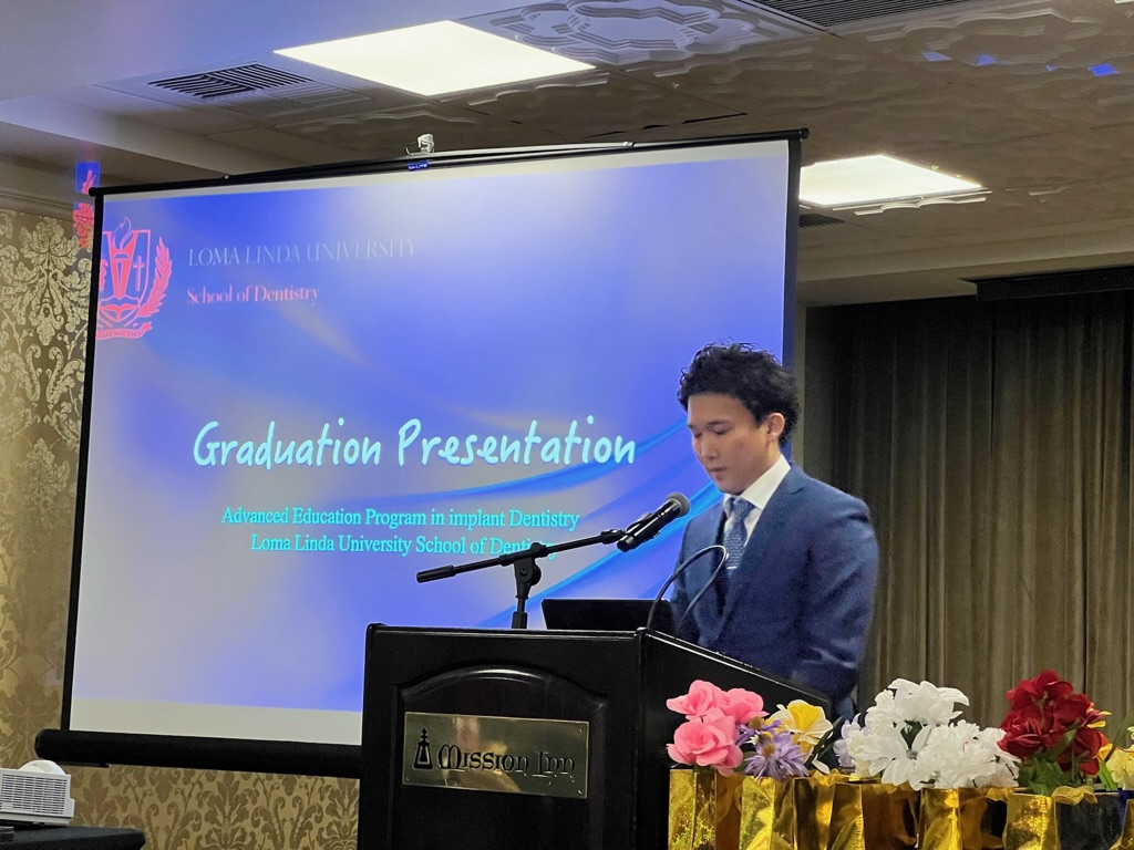 演台でマイクの前に立ち話す著者。背後のスクリーンに Loma Linda University School of Dentistry、Graduation Presentation、Advanced Education Program in implant Dentistry と表示されている